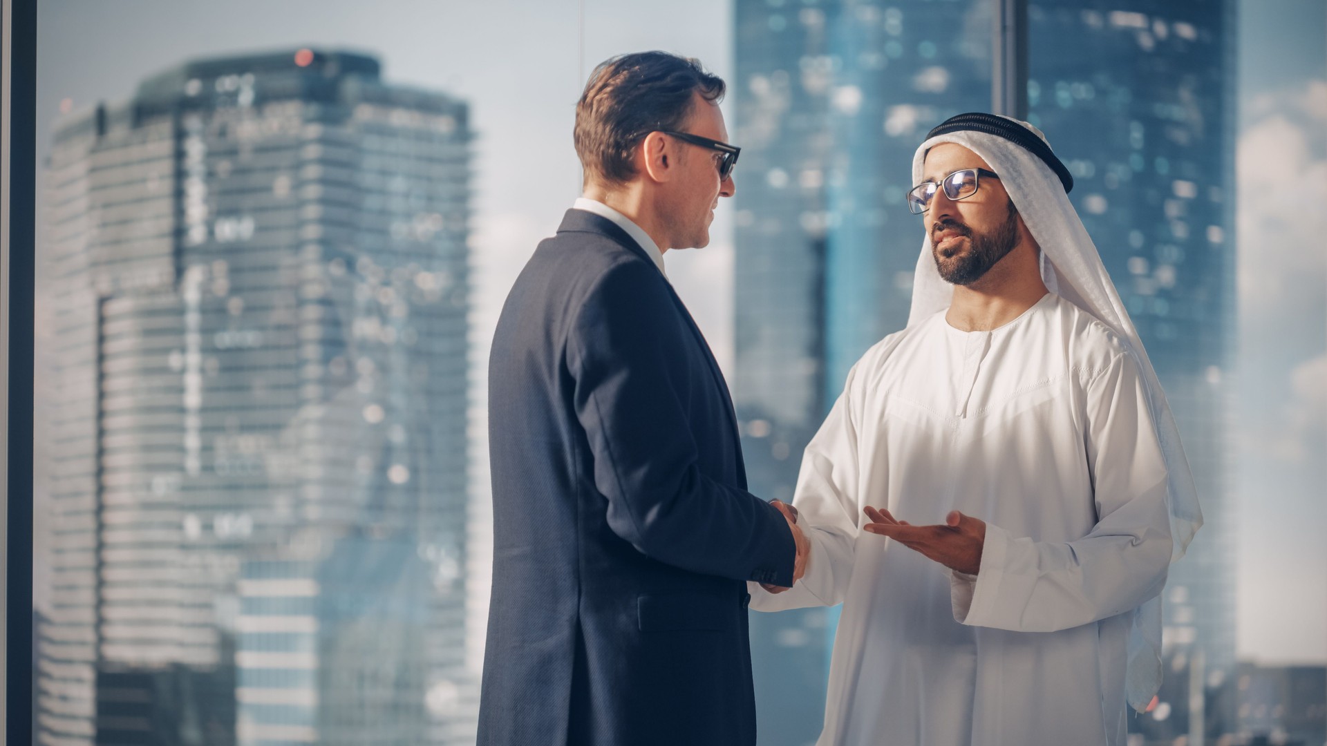 International Operations Manager Meeting Muslim Business Partner in Traditional White Kandura. They're Standing in Modern Office, Shaking Hands. Successful Saudi, Emirati, Arab Businessman Concept.